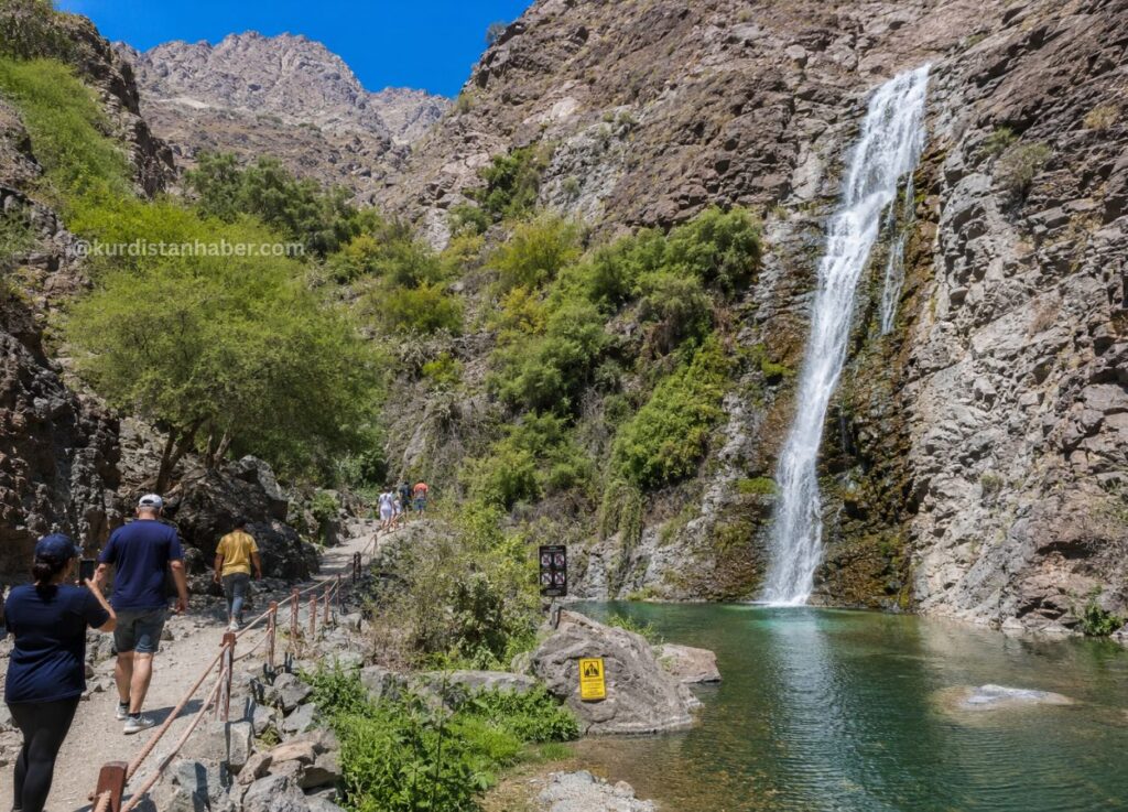 Hikers walk along a rocky trail beside a tall waterfall cascading into a turquoise pool, with cliffs and green trees under a blue sky.